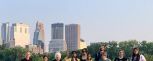 11 people standing in front. Several skyscrapers of Minneapolis can be seen to the left behind the people, with trees on the right. Two women wear black T-shirts with the blue & green Fairtrade logos. The T-shirts say, "the future is fair" There are 1 man and 10 women. A woman from India holds two fingers up as a "peace" sign.