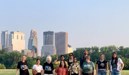 11 people standing in front. Several skyscrapers of Minneapolis can be seen to the left behind the people, with trees on the right. Two women wear black T-shirts with the blue & green Fairtrade logos. The T-shirts say, "the future is fair" There are 1 man and 10 women. A woman from India holds two fingers up as a "peace" sign.
