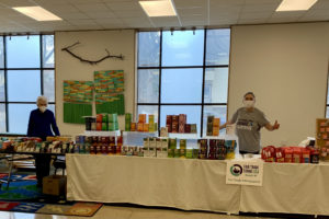 Two women standing behind three tables connected lengthwise, about 30 feet of tables covered with Fair Trade chocolate, coffee, tea, & artisan-made products