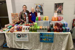 Two women stand behind a table. The table has a white tablecloth, and is covered with Fair Trade products