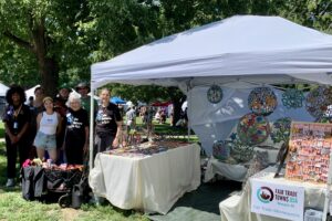 White canopy tent with 3 tables. Bulletin board full of earrings and sign for Fair Trade Minneapolis. To left: 7 people who helped with this booth.