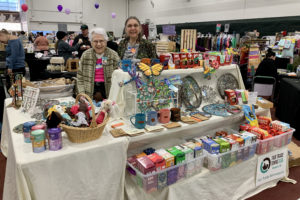 2 women stand behind 2 tables covered with Fair Trade products