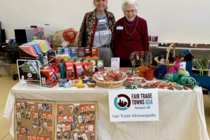 Two women standing behind a table. There's a tablecloth on the table, and bulletin boards containing earrings in front of the table. The table is filled with Fair Trade products: coffee, tea, artisan products, etc.