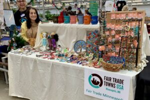 Two women stand at the left side behind a table. The table has a bench on top, with an ivory-colored sheet over all of it. On this, 100s of artisan-made Fair Trade products are displayed - ceramic mugs, dolls, holiday ornaments, bracelets, metal wall art, necklaces & earrings