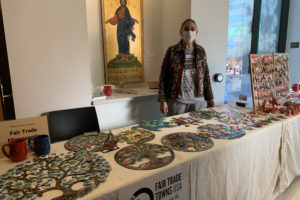 a woman stands behind two long tables covered with white tablecloths & artisan-made Fair Trade products: metal wall art, coffee mugs, earrings, & holiday ornaments.