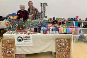 Two women stand behind a table covered with Fair Trade products: chocolate, coffee, tea, artisan products from Haiti, etc.