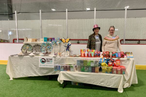two women stand behind a table. The table has an ivory-colored table cloth. On the table: Several kinds of Fair Trade chocolate including Tony's Chocoloney, Equal Exchange, and Singing Rooster. In front of the table: a bench holding two bins of dozens of kinds of Fair Trade tea, and one bin of Fair Trade coffee.