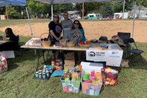 Three white women standing behind a table under a canopy, outside, in a city park. On table: artisan Fair Trade products. Under table: boxes of Fair Trade tea