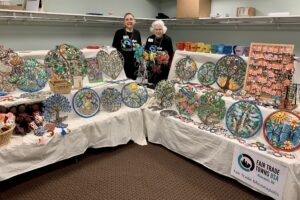 two women stand between two long tables. With a bench on each table, and a bench beneath, the tables display metal wall art, ceramic mugs, and jewelry made in Haiti
