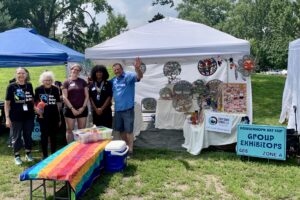 five people standing to the left of a white canopy booth. 3 tables covered with ivory-white sheets support metal art from Haiti. Right: A bulletin board full of earrings. Left: 2 benches draped with a rainbow-striped beach towel and a bin of boxes of free Fair Trade tea, with a blue cooler full of free Fair Trade chocolate.