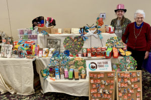 2 women stand to the right of a large display of Fair Trade products, made by 2 tables. Under ivory colored tablecloths, a bench on the main table, and a bench in front of it, make the display