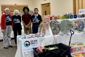 4 people stand in a booth. Bracelets, earrings, ceramic mugs, dolls in a wagon, boxes of tea, and metal art are on and near several tables