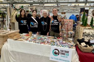 4 women stand behind a table in a marketplace - in a greenhouse! On the table: greeting cards to left, holiday ornaments, and bracelets.
