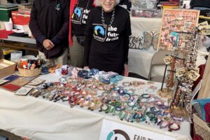 3 people stand behind a table in an indoor marketplace. On the table: freeing cards, a whole rainbow of bracelets, necklaces on standers. Behind: earrings and metal art.