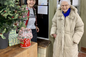 Next to a Christmas tree, near the door to a store at night, two women stand near the window. Between them, a Fair Trade Award has been posted.