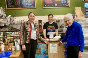 One Native woman stands behind the counter in a cafe. Two white women stand in front of the counter, pointing at the Fair Trade Award posted just beneath the counter